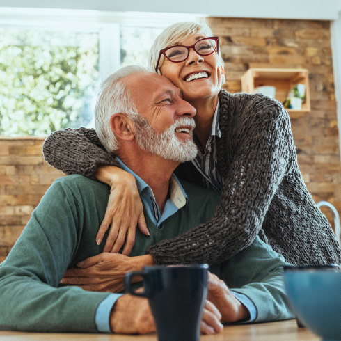 A mature couple smiling and embracing at a wood table in a kitchen.
