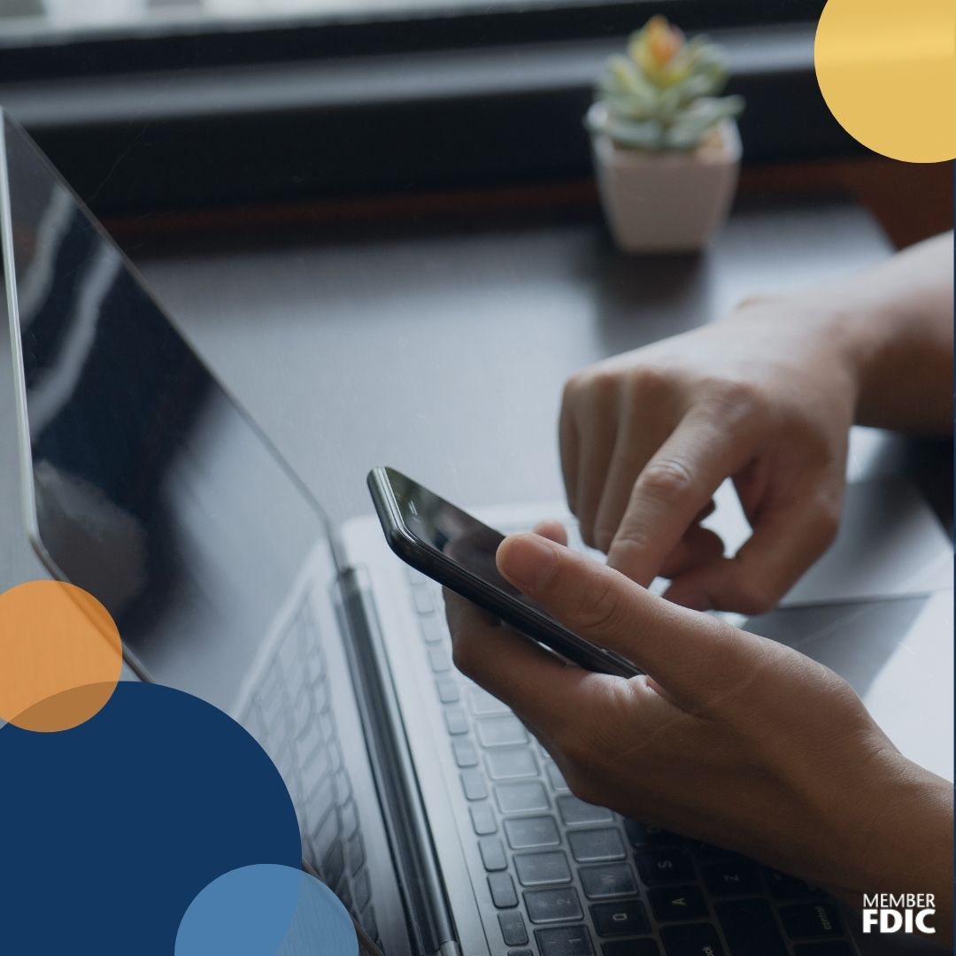 A picture of a person's hands holding their phone. The person's laptop is in the background of the picture. There is nothing shown on the phone or computer screens.