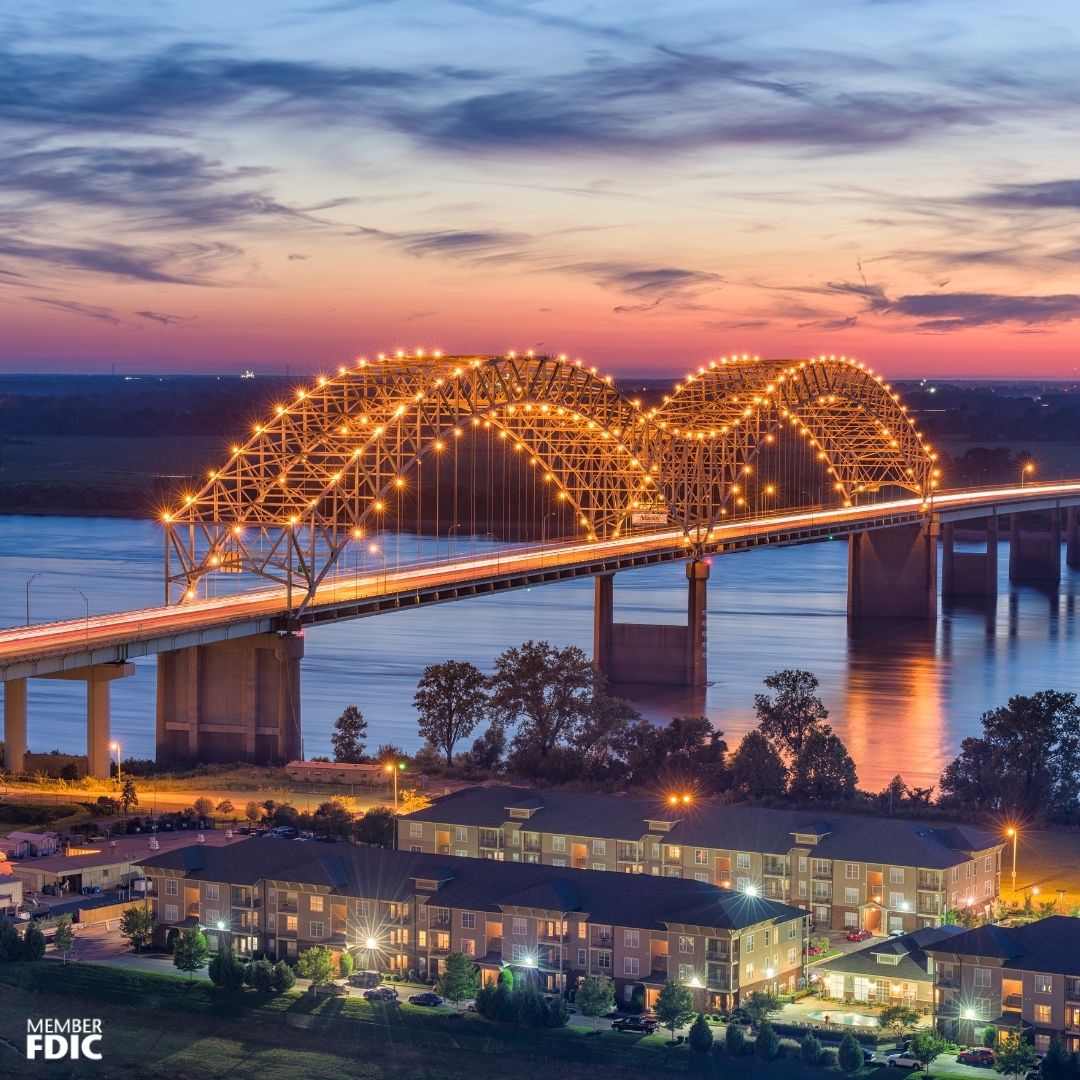 A picture of a lit up bridge over the river with houses nearby. A colorful sunrise is in the background of the image. 