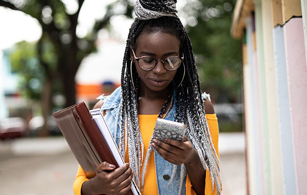 Young woman holding school supplies and looking at her phone