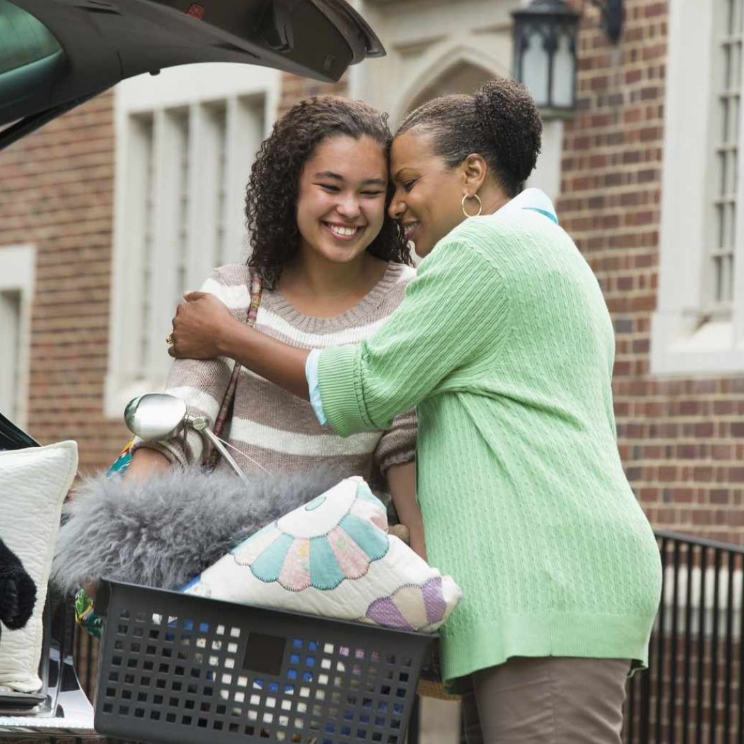 A mother hugging her daughter as she puts a box of things into the car. The daughter is going off to college and packing her things to leave. The mother and daughter are smiling.