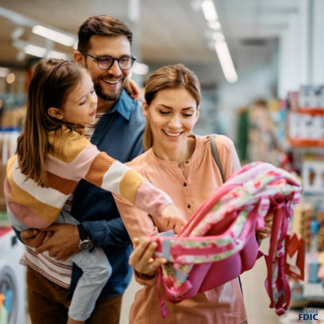 family of three shopping for a backpack for their daughter. the father is holding the daughter, who is pointing to a backpack she likes. the family is happy.