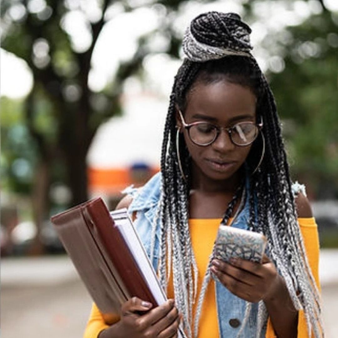 A picture of a woman with glasses looking down at her phone in one hand while the other hand is holding onto books.